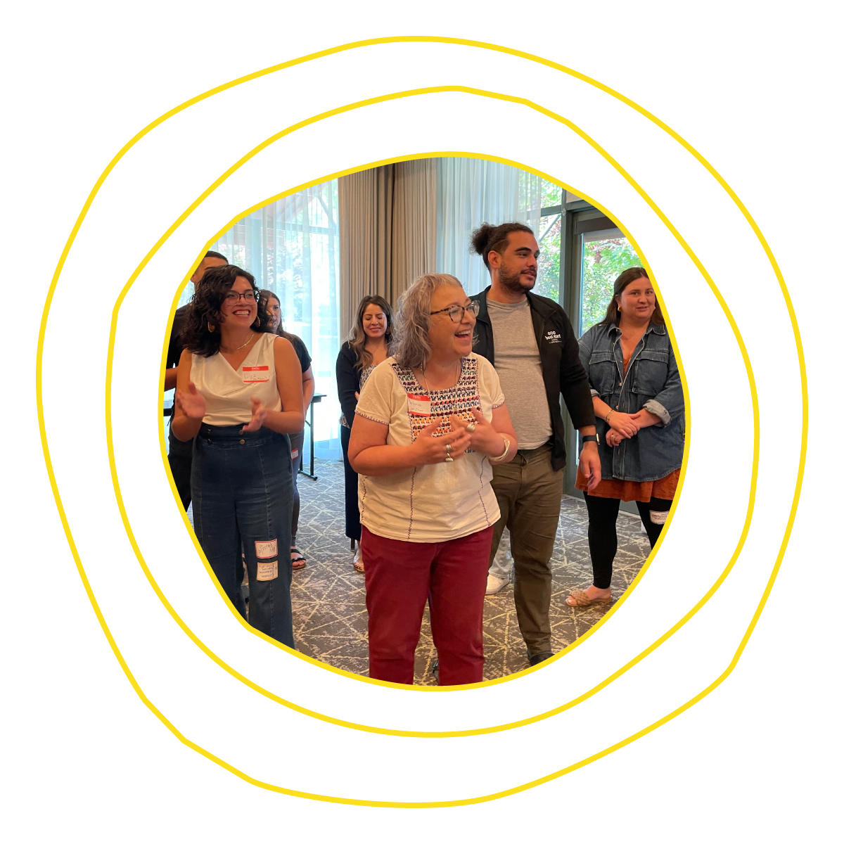 Within a room illuminated by large windows, a diverse group of people are gathered. Their collective focus is directed to something to the left that is both enthralling and joyous.