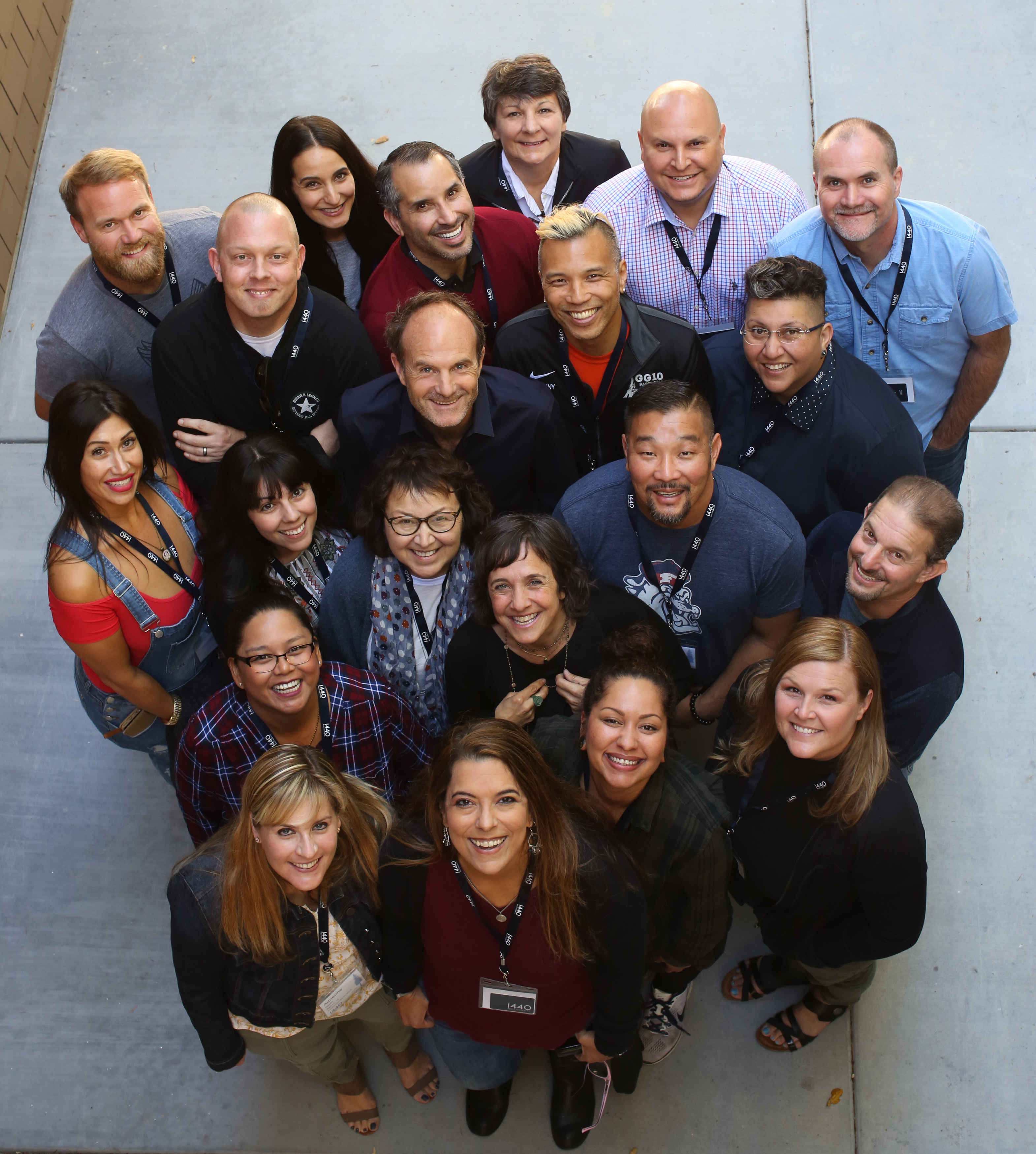 A diverse group of individuals wearing lanyards are standing close together, smiling and gazing up towards the photographer for a group photo.